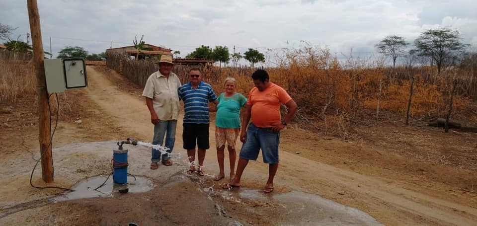 Quadro pessoas em frente a um equipamento jorrando água em uma estrada de terra. Ao fundo cercas, mata seca e casas.