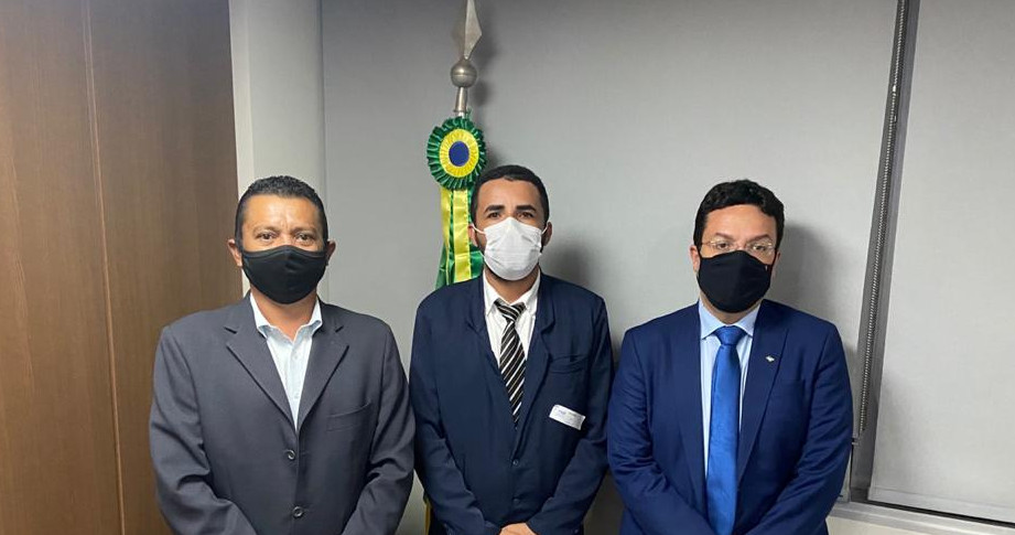 Três homens, lado a lado, trajando terno e usando máscara