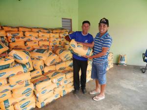 Duas pessoa segurando um saco de milho e ao lado tem v&aacute;rios sacos empilhados no ch&atilde;o.