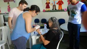 Foto com dois homens em p&eacute; e uma mulher fazendo teste de glicemia em uma senhora sentada encostada numa mesa.